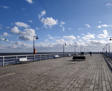 Rozległy drewniane molo  z białymi balustradami zachęca do spaceru nad Zatoką Pucką. Słoneczny kadr prezentuje popularne molo w Jastarni i otwartą przestrzeń nad wodą.