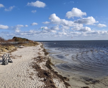 Piaszczysty brzeg, drewniane schody i płytka woda pokazują naturalny krajobraz Jastarni od strony Zatoki Puckiej. Wiosenne chmury podkreślają przestrzeń P&oacute;łwyspu Helskiego.